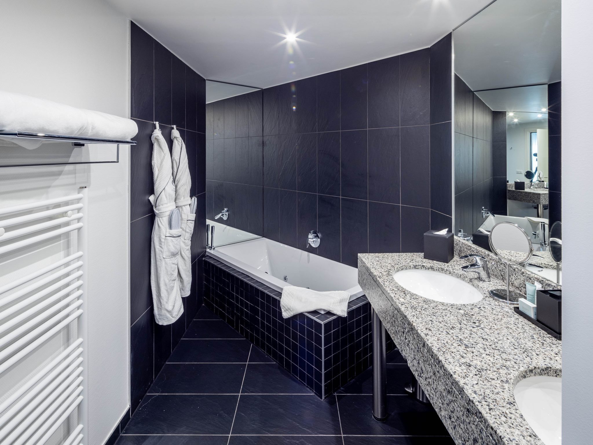 Un baño moderno con bañera, dos lavamanos, baldosas oscuras y un espejo grande.