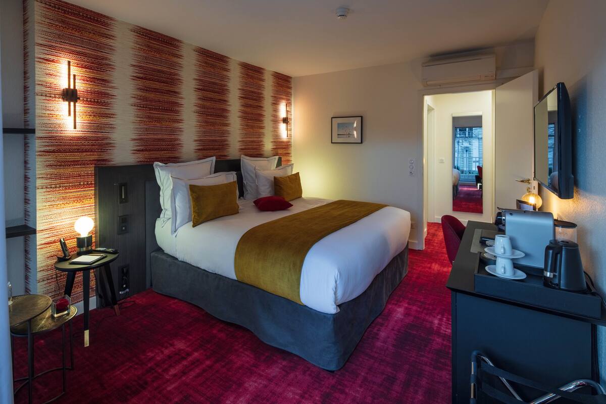A hotel room with a large bed, a desk with a coffee maker, a red carpet, and a wall with a textured pattern.
