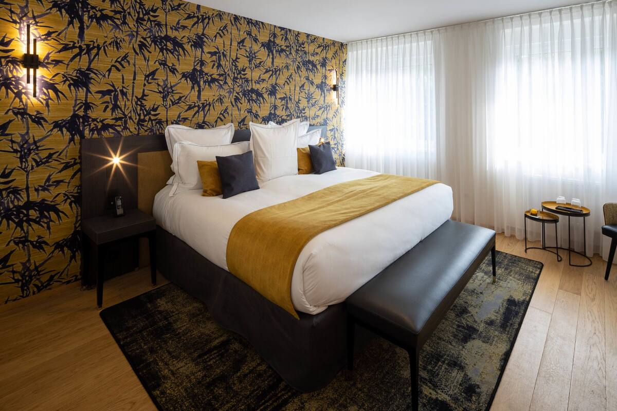 A modern hotel room with a large bed, a bedside table, a bench, and a small table with a chair.