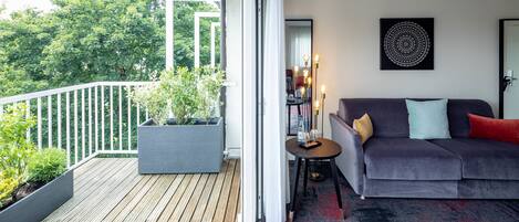 Un salón moderno con un sofá gris, una mesita auxiliar y un ventanal grande que da a una terraza de madera y follaje.