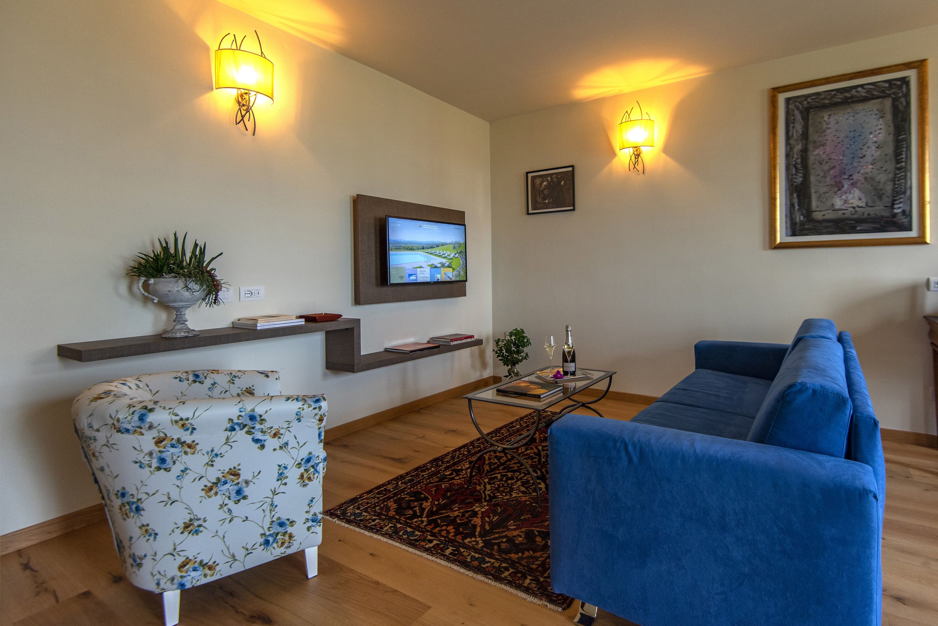 Una sala de estar con un sofá azul, un sillón floral, un televisor de pantalla plana, lámparas fijadas a la pared y un cuadro enmarcado.