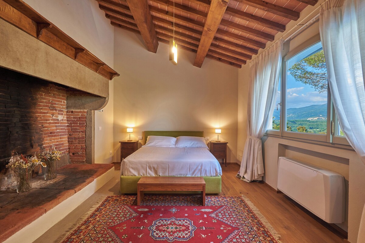 Un dormitorio con chimenea de ladrillo, una cama verde, un tapete rojo con estampado y vistas al exterior a través de una ventana con cortinas blancas.