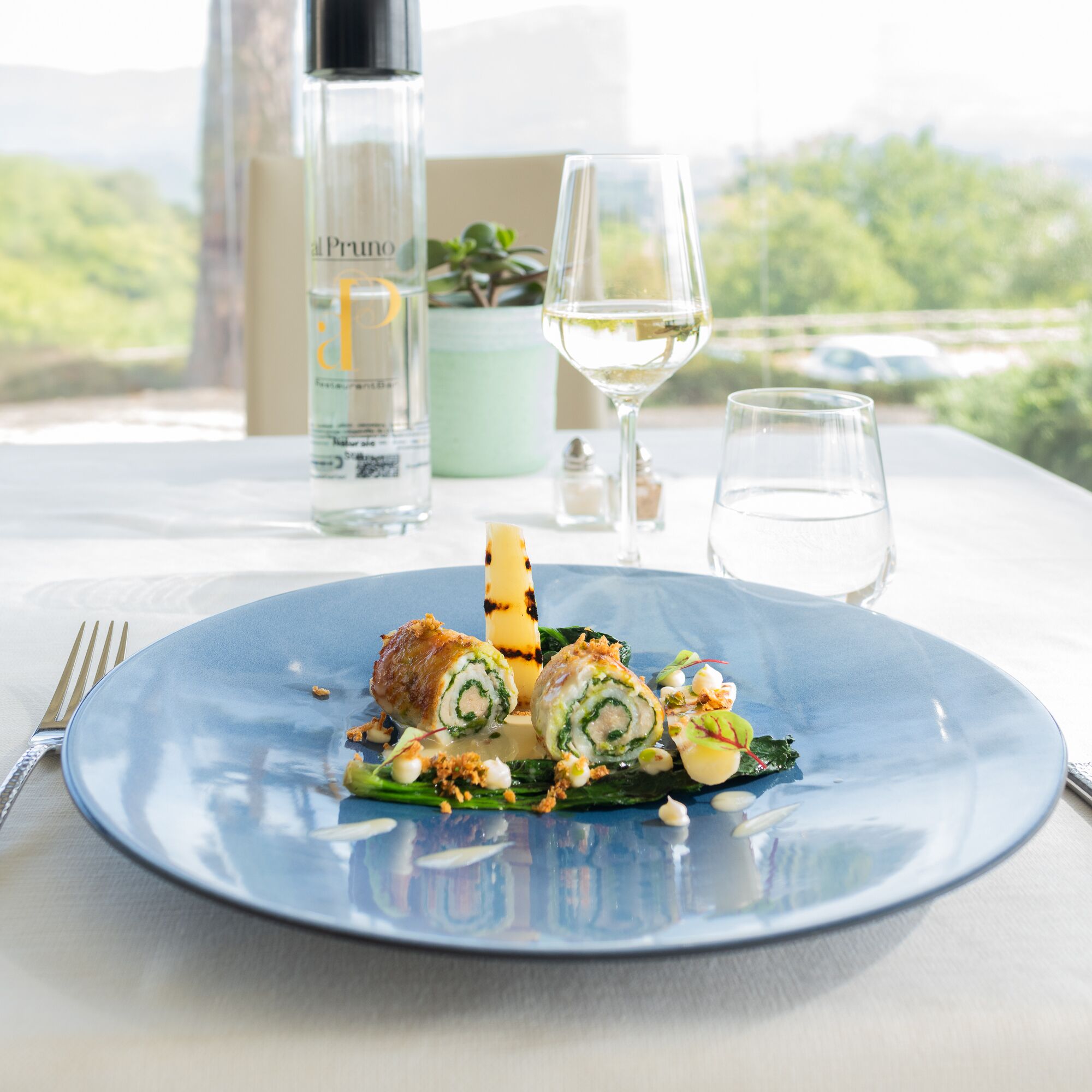 Un piatto blu con un rotolo di cibo, un bicchiere di vino bianco e una bottiglia etichettata 'Pruno' su un tavolo con vista sul verde.