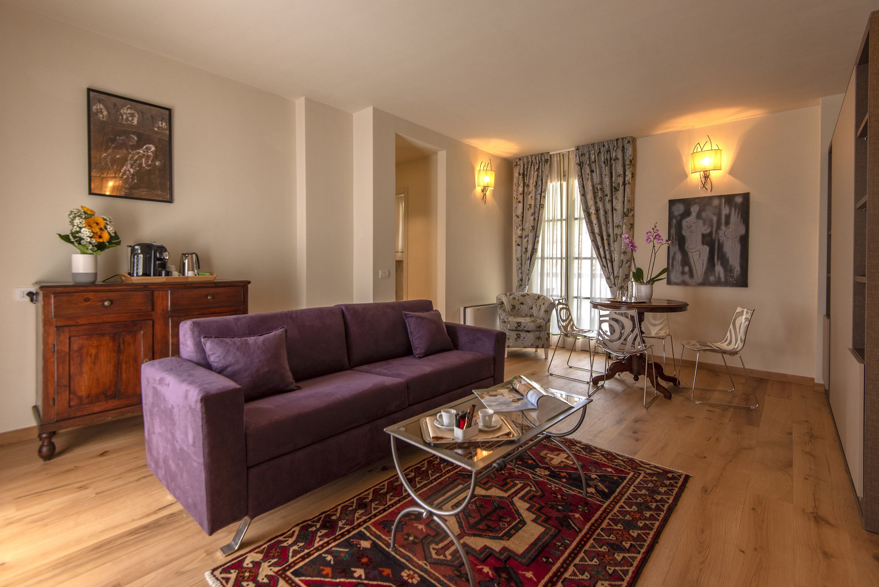 A living room with a purple sofa, a glass coffee table, a patterned rug, a wooden cabinet, and a floral vase.