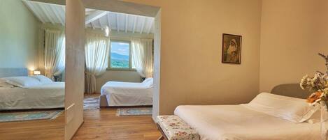 A room with two beds, a wooden floor, a large window with curtains, and a painting on the wall.