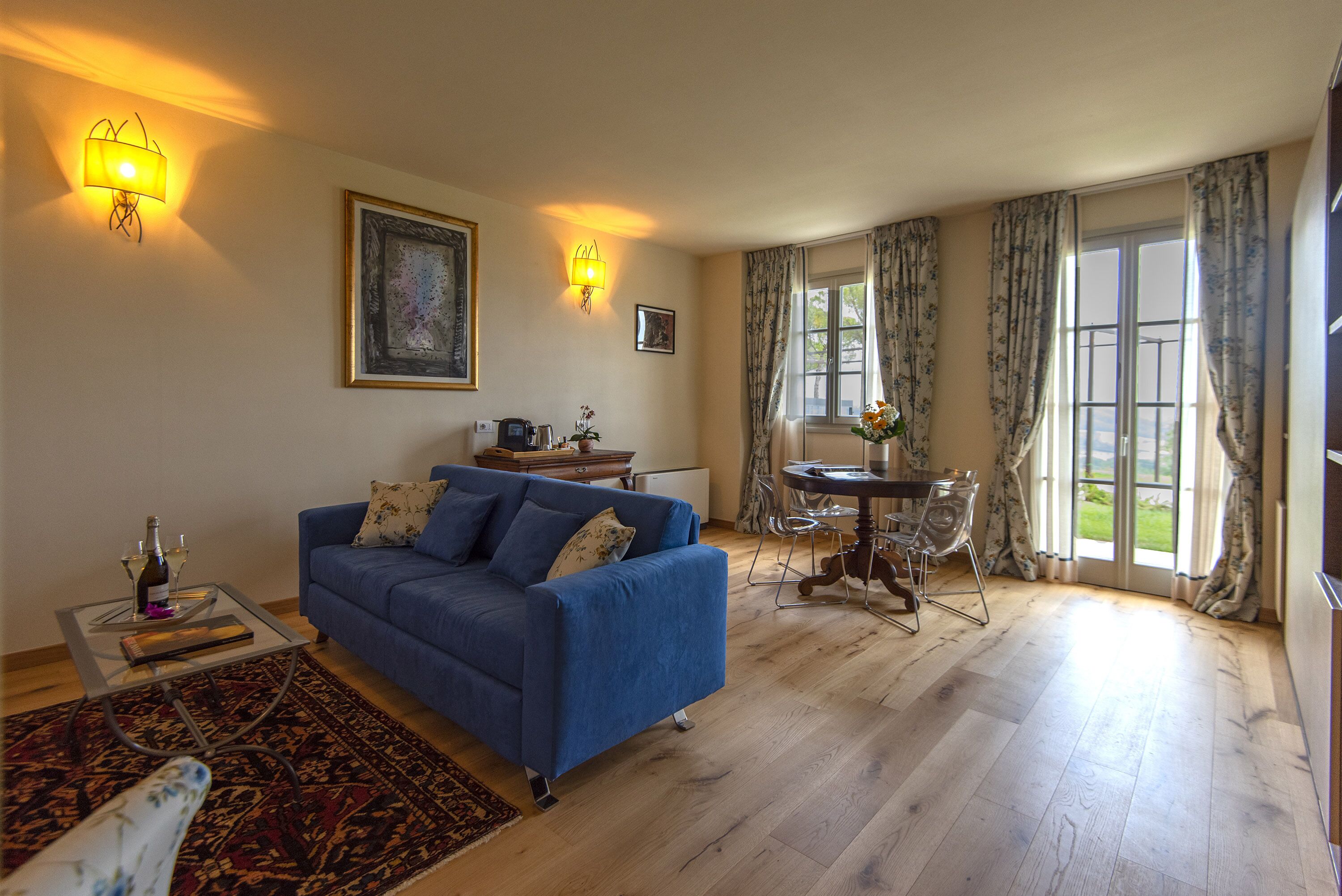 Sala de estar com um sofá azul, uma mesa de centro de vidro, uma mesa de jantar com cadeiras e vista para o exterior através de grandes janelas.