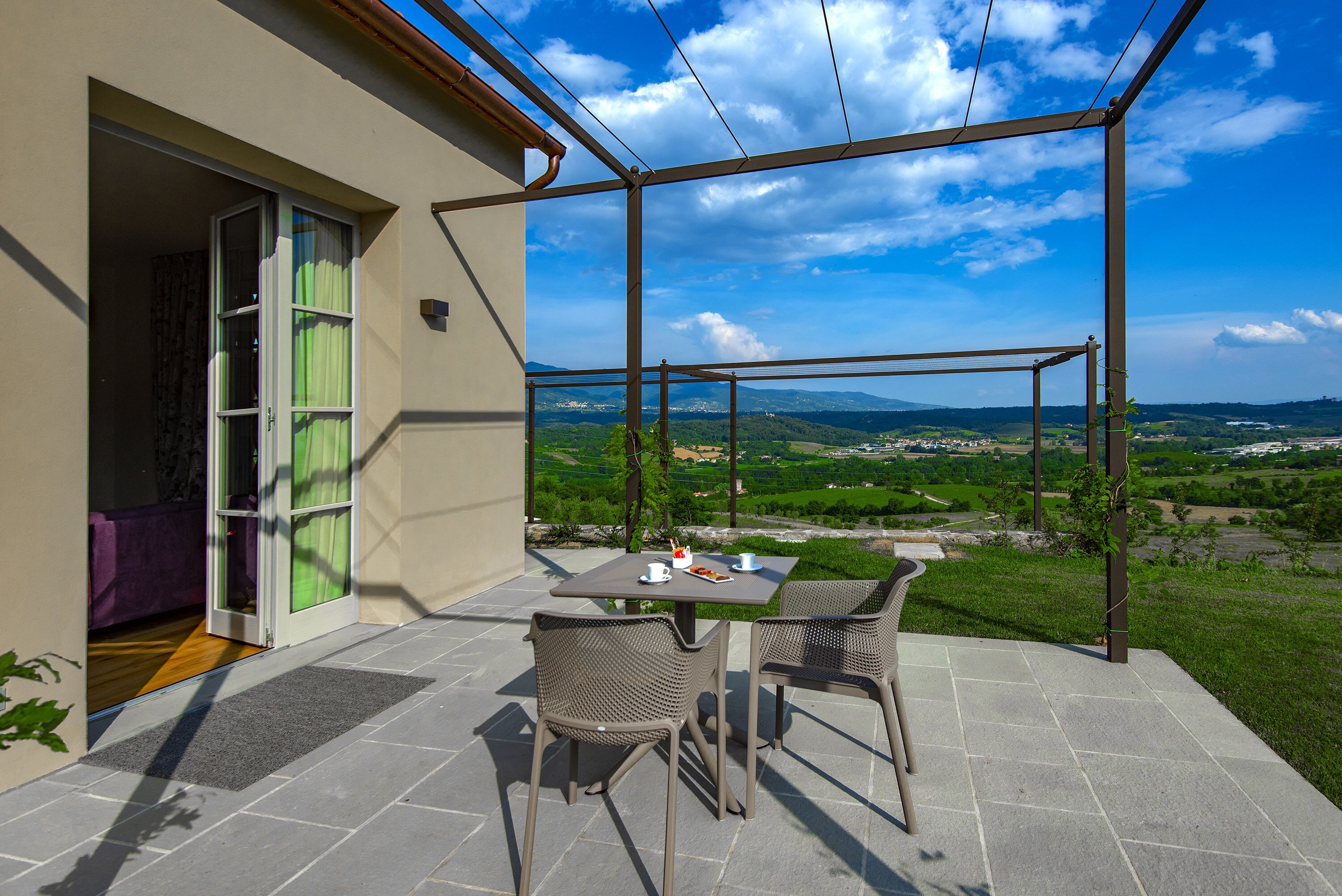 Une terrasse extérieure avec une table et des chaises, une vue sur un paysage verdoyant et l’entrée d’un bâtiment.