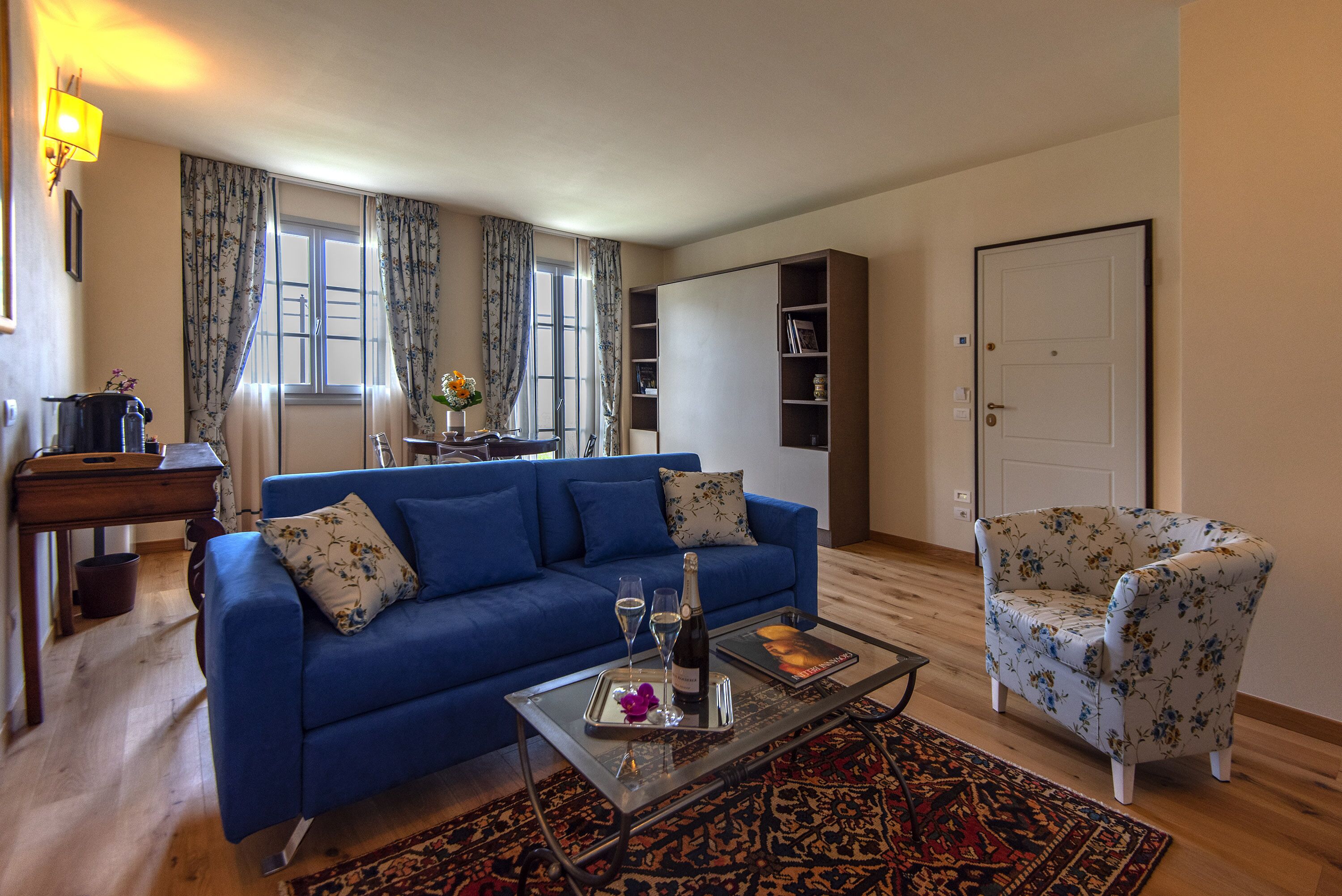 A living room with a blue sofa, a floral armchair, a glass coffee table with a tray, and a bookshelf.