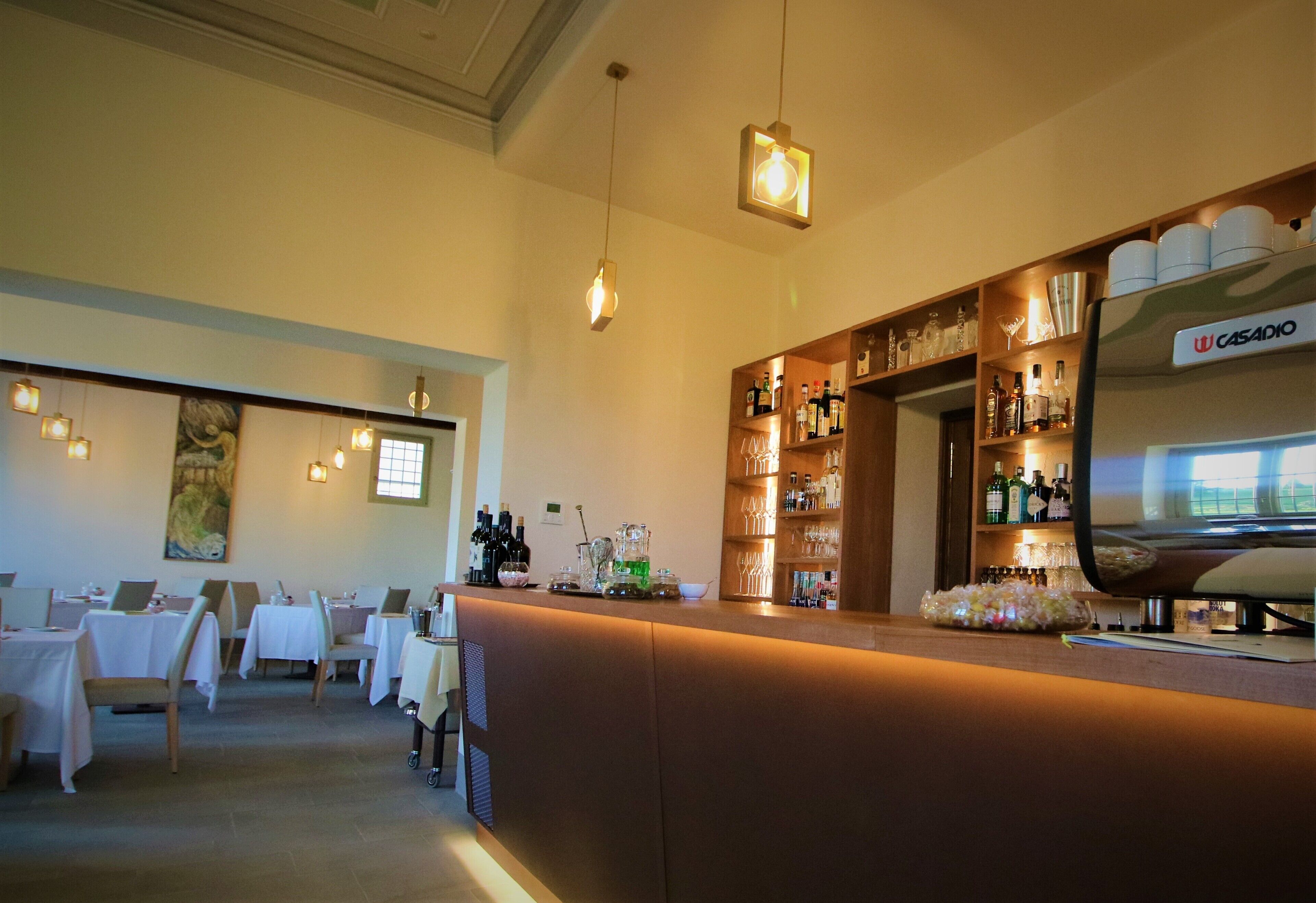 A bar area with a counter, shelves of bottles, and a television screen. Tables and chairs are arranged in the background.