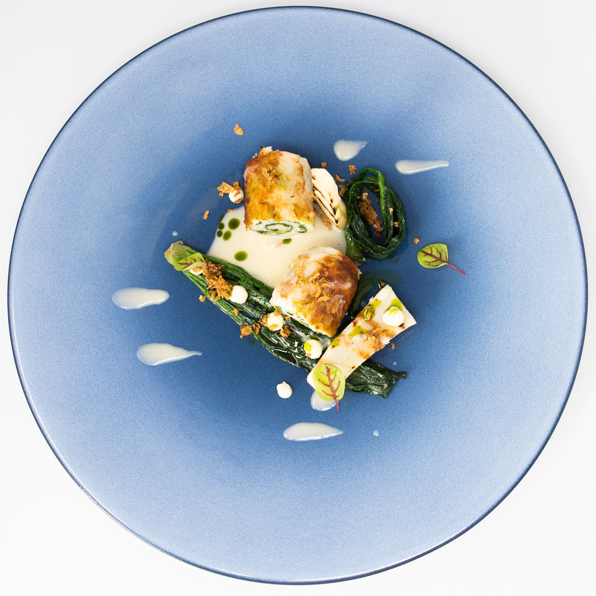 A blue plate with a dish consisting of a piece of breaded meat, green vegetables, and a creamy sauce.