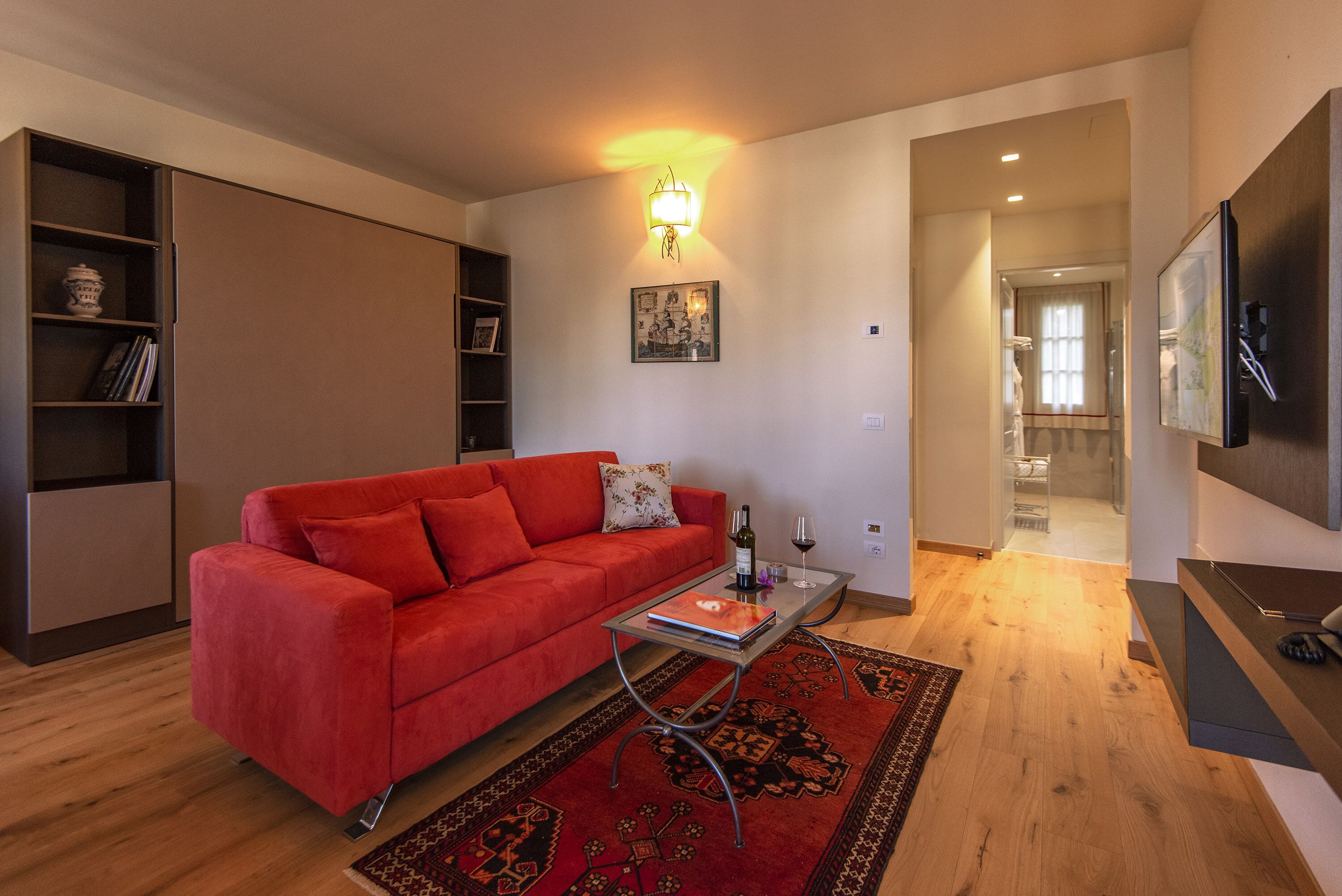 Un salon avec un canapé rouge, une table basse en verre, un tapis à motifs, un luminaire fixé au mur et un téléviseur placé à droite.