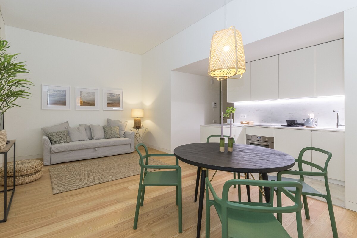 A modern living room with a grey sofa, green chairs, a round dining table, and a kitchen area with white cabinets and a sink.