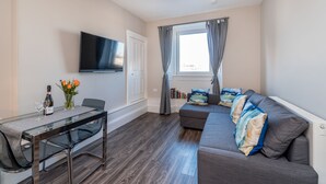 A modern living room with a glass dining table, a sofa with patterned cushions, a flat-screen TV mounted on the wall, and a window with curtains. - Silver Lining Charming Meadowbank Flat (Edinburgh)