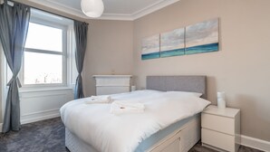 A neatly made bed with white linens and towels, a bedside table with a lamp, and a painting of a seascape on the wall. - Silver Lining Charming Meadowbank Flat (Edinburgh)