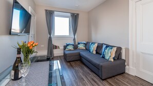 A modern living room with a grey sectional sofa, a flat-screen TV, a glass coffee table, and a decorative vase with flowers. - Silver Lining Charming Meadowbank Flat (Edinburgh)
