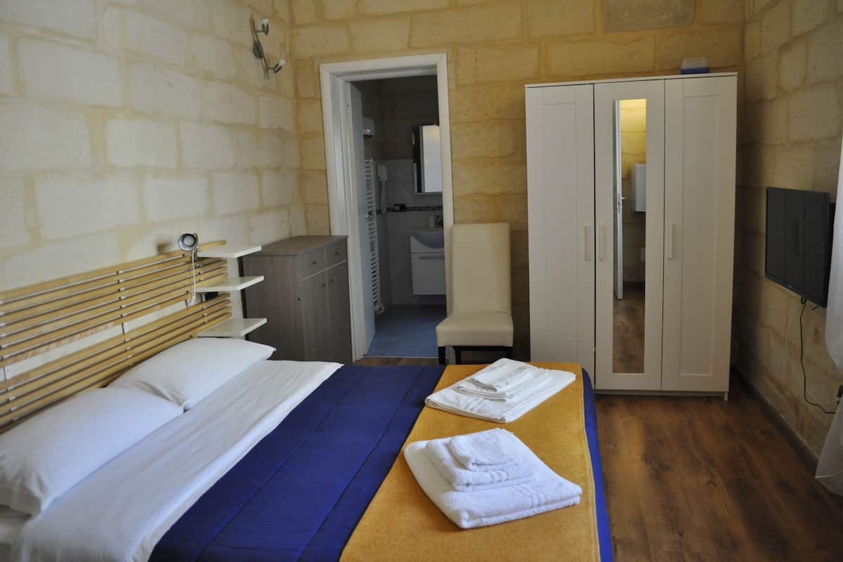 Une chambre d’hôtel avec un lit, une tête de lit en bois, une couvre-lit bleue, une table en bois avec des serviettes, une armoire et une salle de bain visible à travers une porte ouverte.