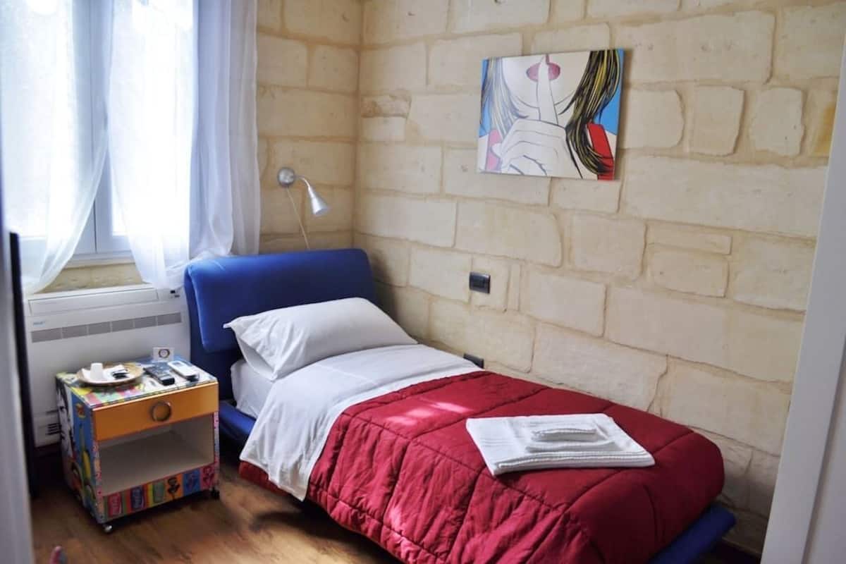 Une petite chambre avec un lit simple, une couvre-lit rouge, un oreiller blanc, une tête de lit bleue, une lampe de chevet, une petite table avec un meuble de rangement coloré et un tableau abstrait accroché au mur.