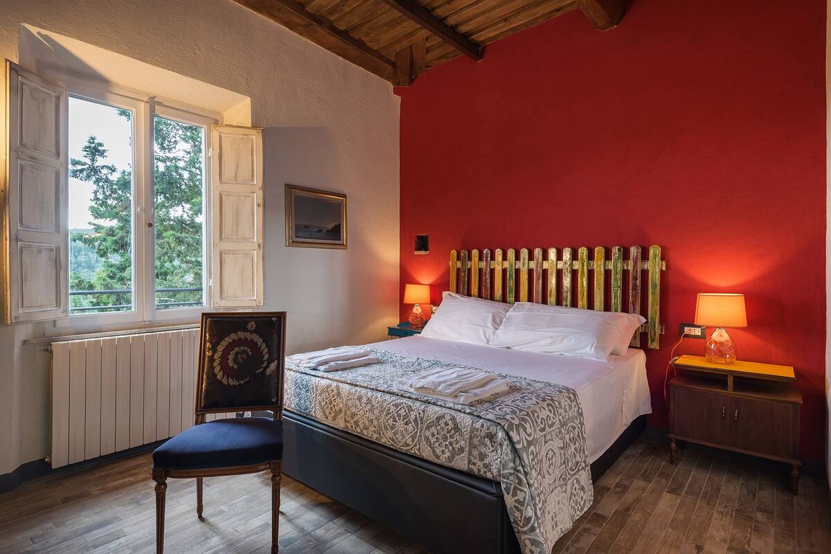 Ein Schlafzimmer mit einem Bett, zwei Nachttischlampen, einem Sessel, einem Fenster mit Blick auf Bäume und einer Heizung.