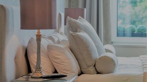A neatly made bed with white bedding and beige pillows, a bedside table with a lamp and remote control, and a window with a view of greenery. - Cults Hotel (Aberdeen)