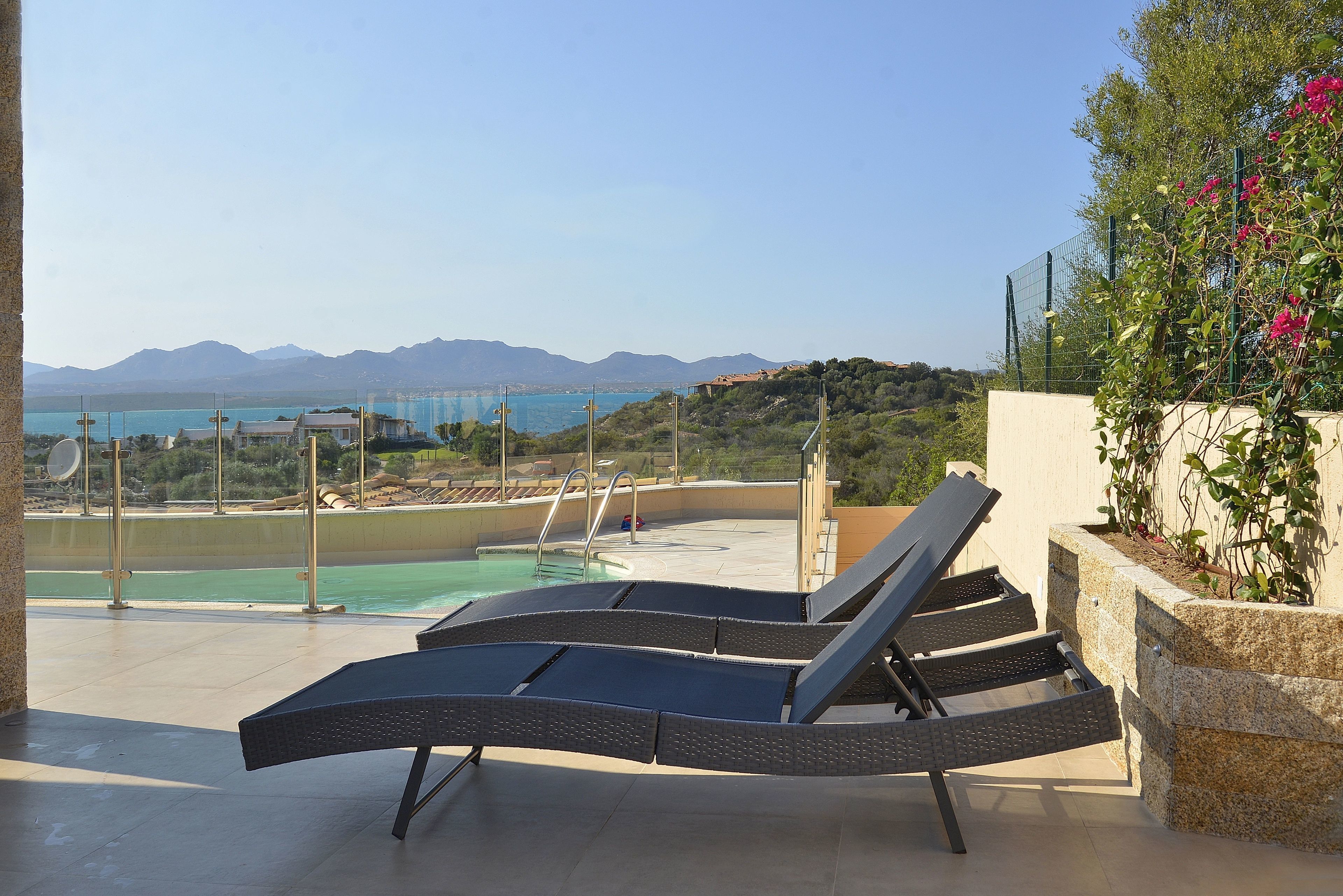 A poolside area with lounge chairs, a pool, and a view of mountains and a body of water.