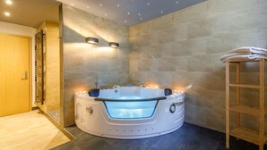 A modern bathroom with a central hot tub, a wooden shelf with towels, and a stone wall.