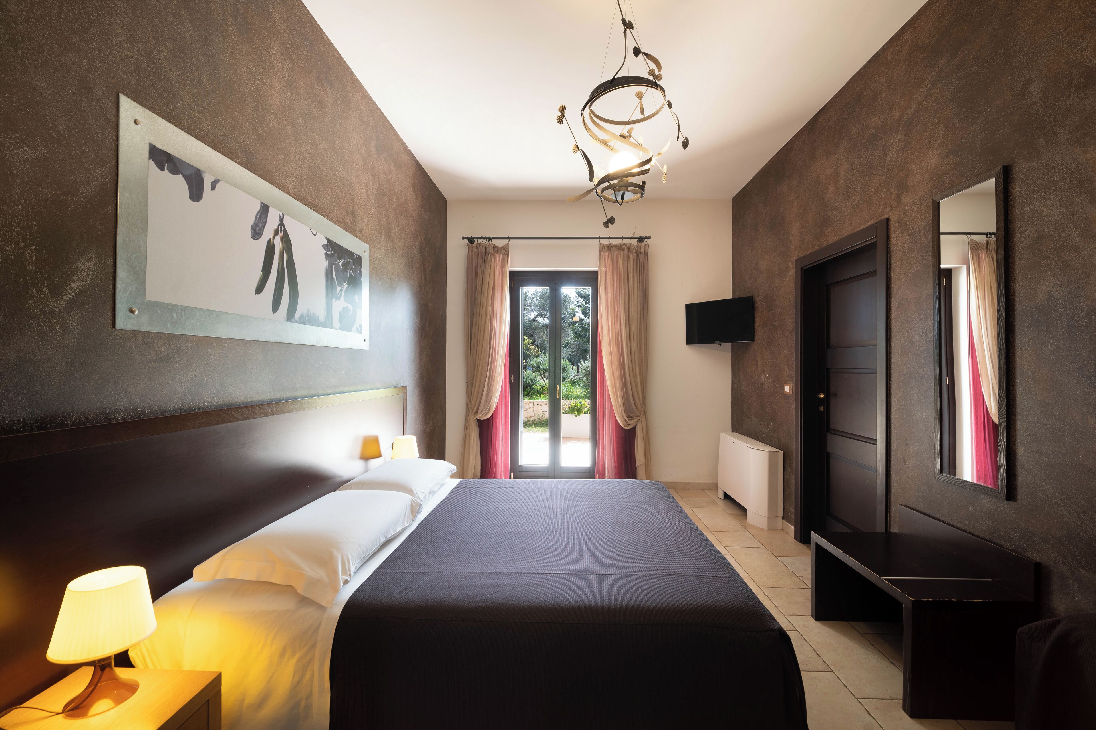 A modern hotel room with a large bed, a bedside lamp, a flat-screen TV, and a view of the outdoors through a sliding door.