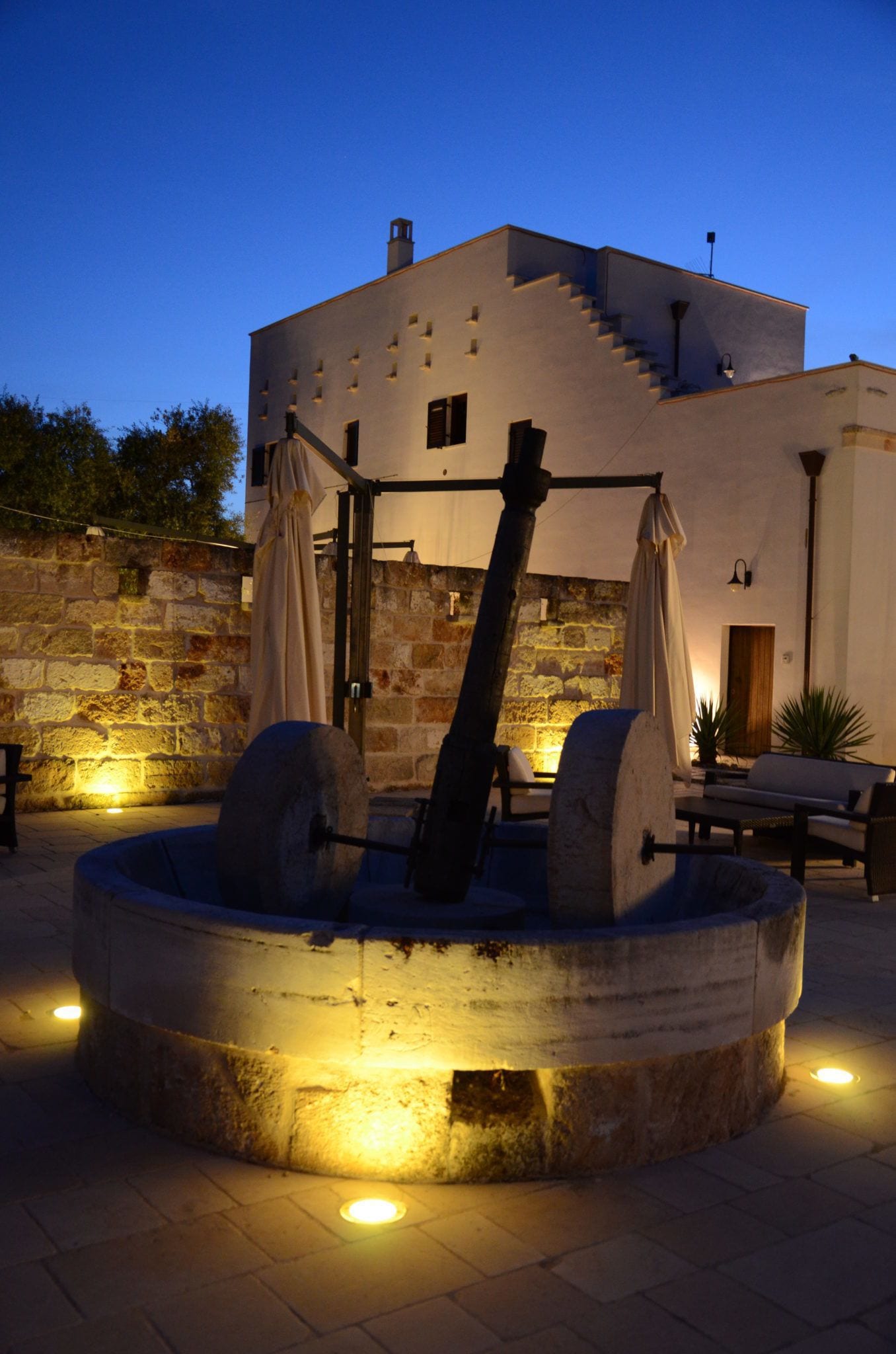 A stone well with a wooden beam in the center, surrounded by a stone wall and a white building with a staircase.