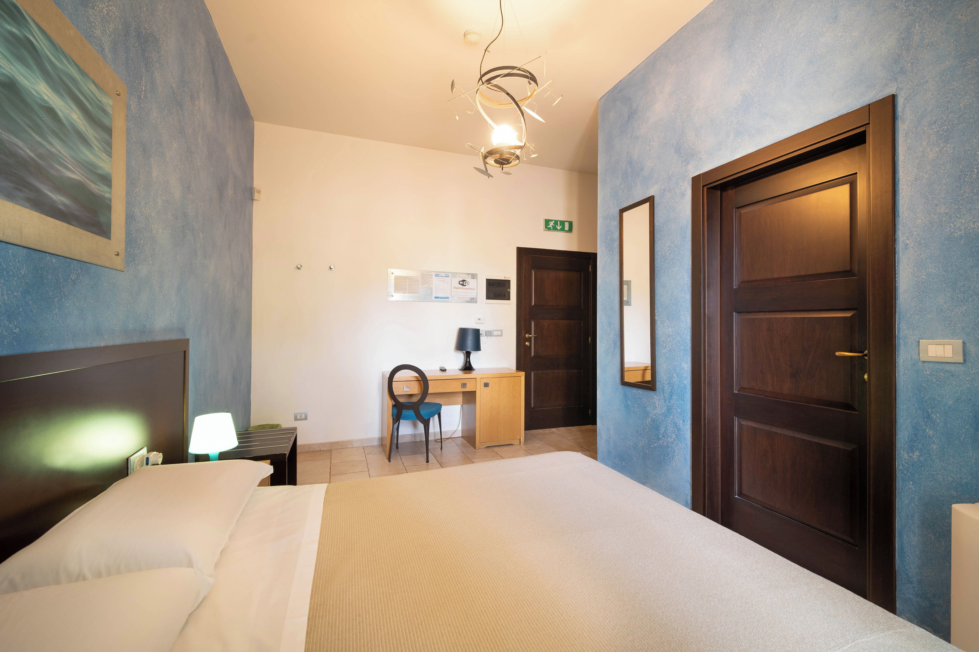 A hotel room with a bed, desk, chair, lamp, and a door.