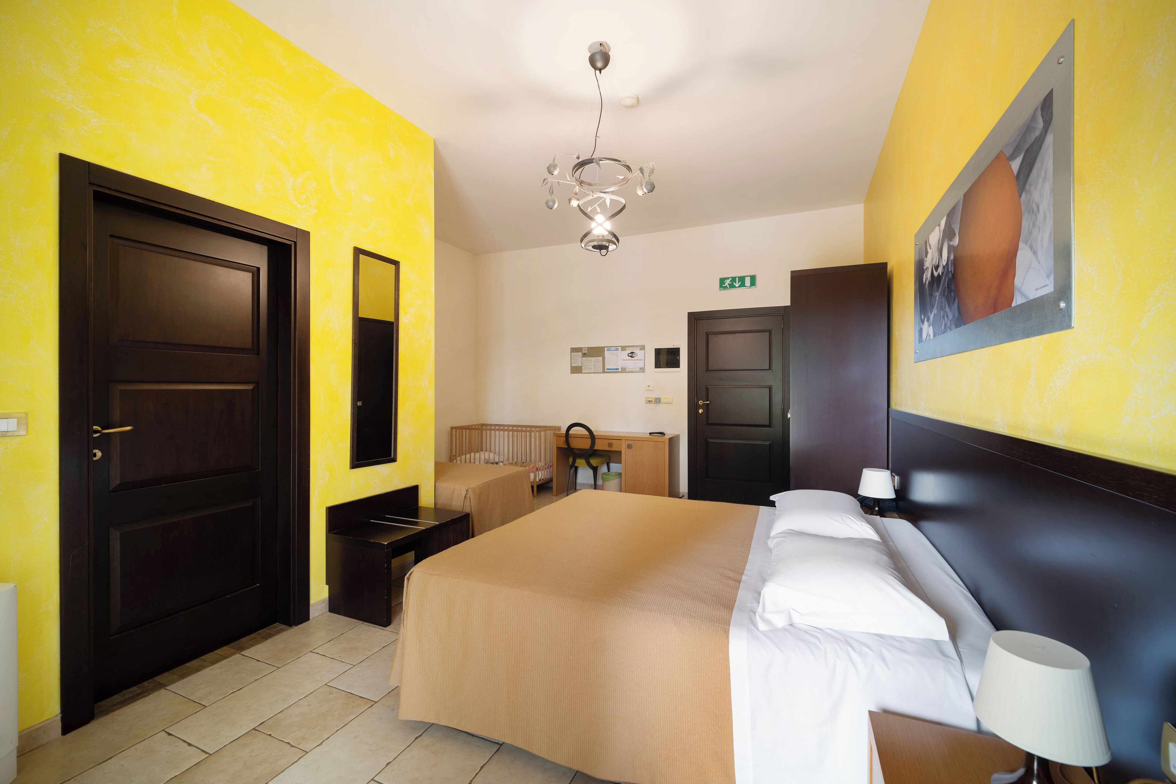 A hotel room with a large bed, a desk, a chair, a crib, and a ceiling light.