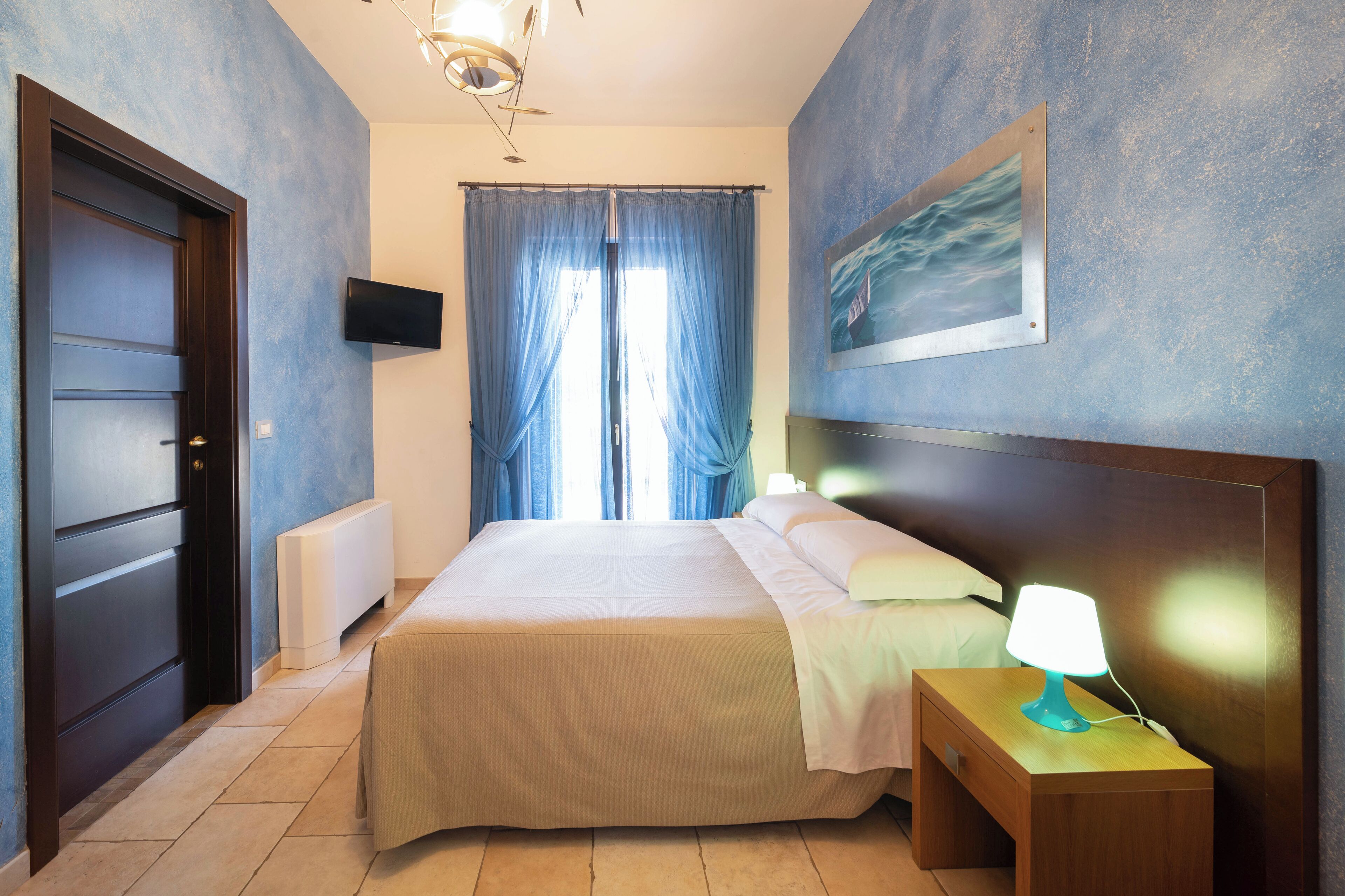 A bedroom with a bed, a nightstand with a lamp, a TV on the wall, and a painting.