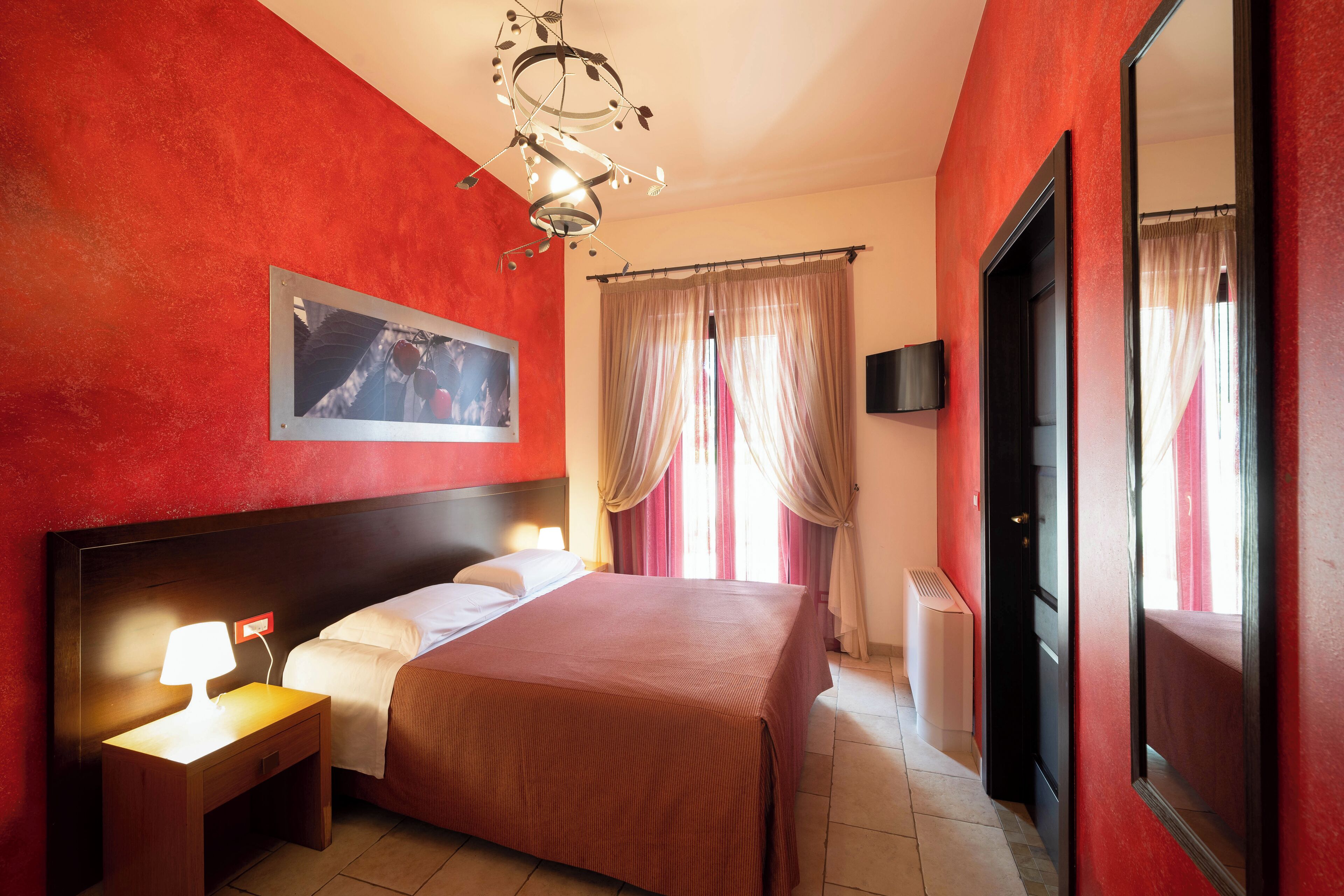 Una habitación de hotel con una cama grande, una mesita de noche con lámpara, un televisor en la pared y una ventana con cortinas.