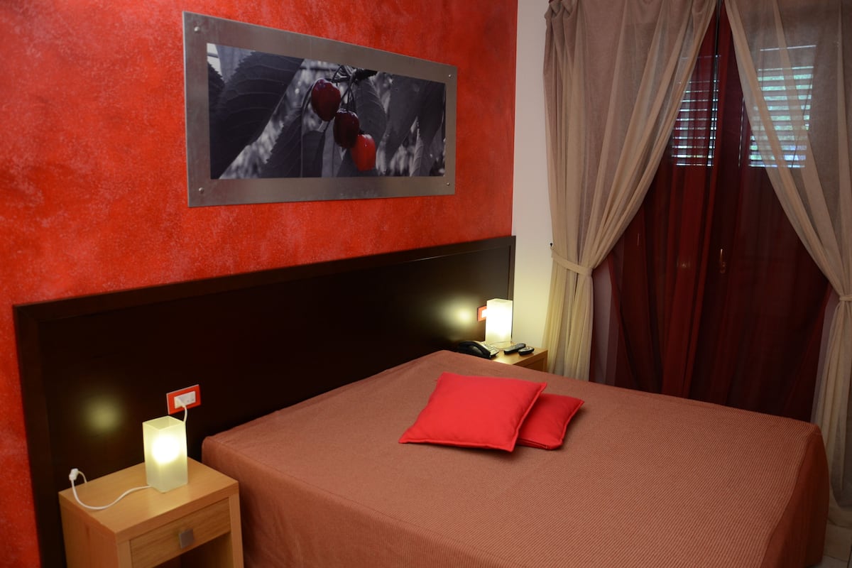 Ein Hotelzimmer mit roter Wand, einem gerahmten Bild, einer Nachttischlampe, einem Telefon und einem roten Kissen auf dem Bett.