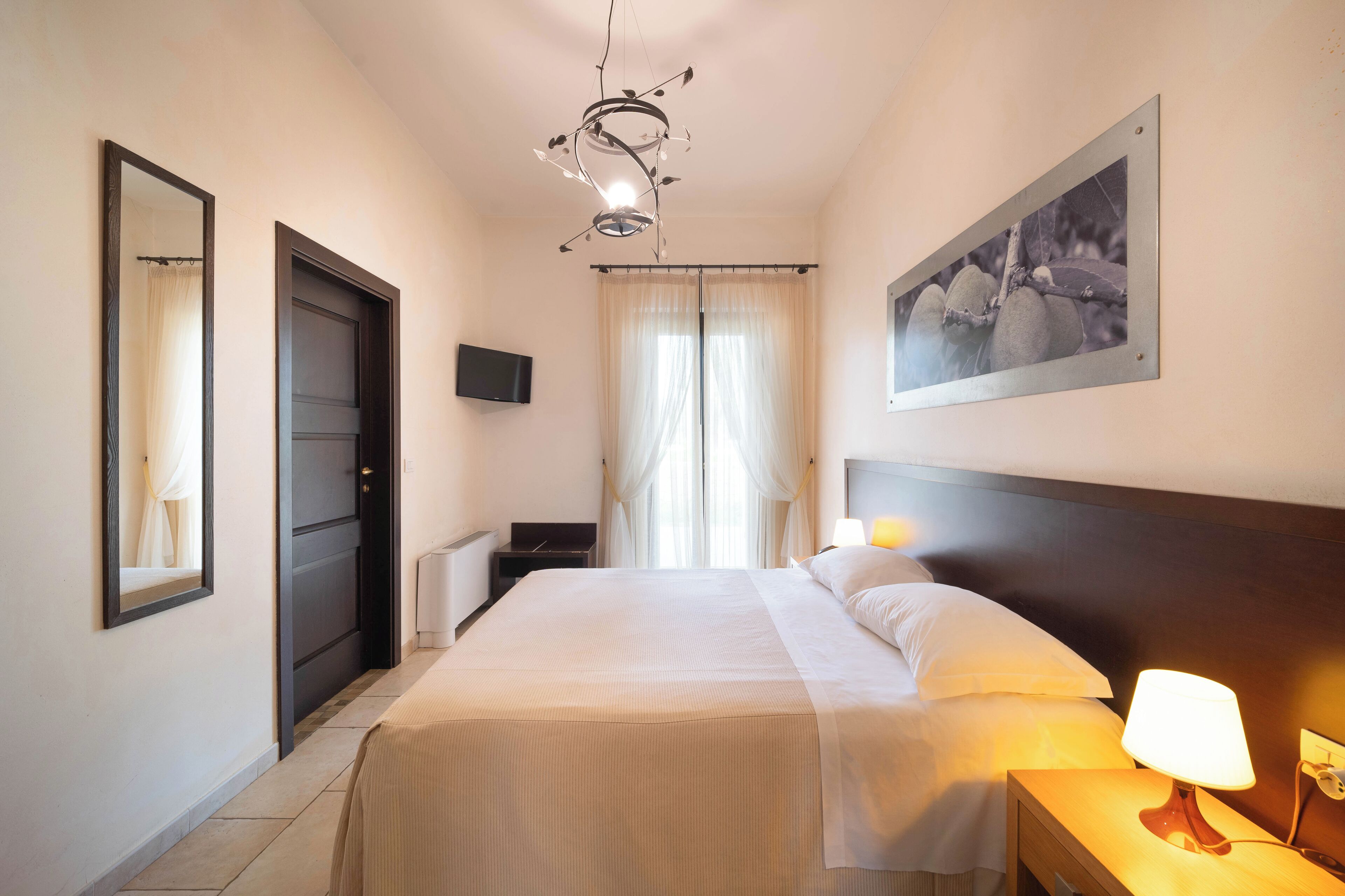 A hotel room with a bed, bedside lamp, a painting on the wall, a TV on the wall, and a window with curtains.