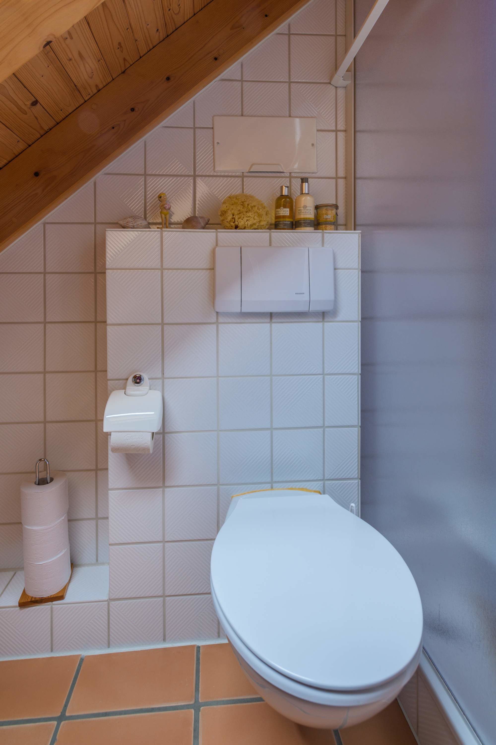 A small bathroom with a white toilet, a paper towel dispenser, and a shelf with toiletries.