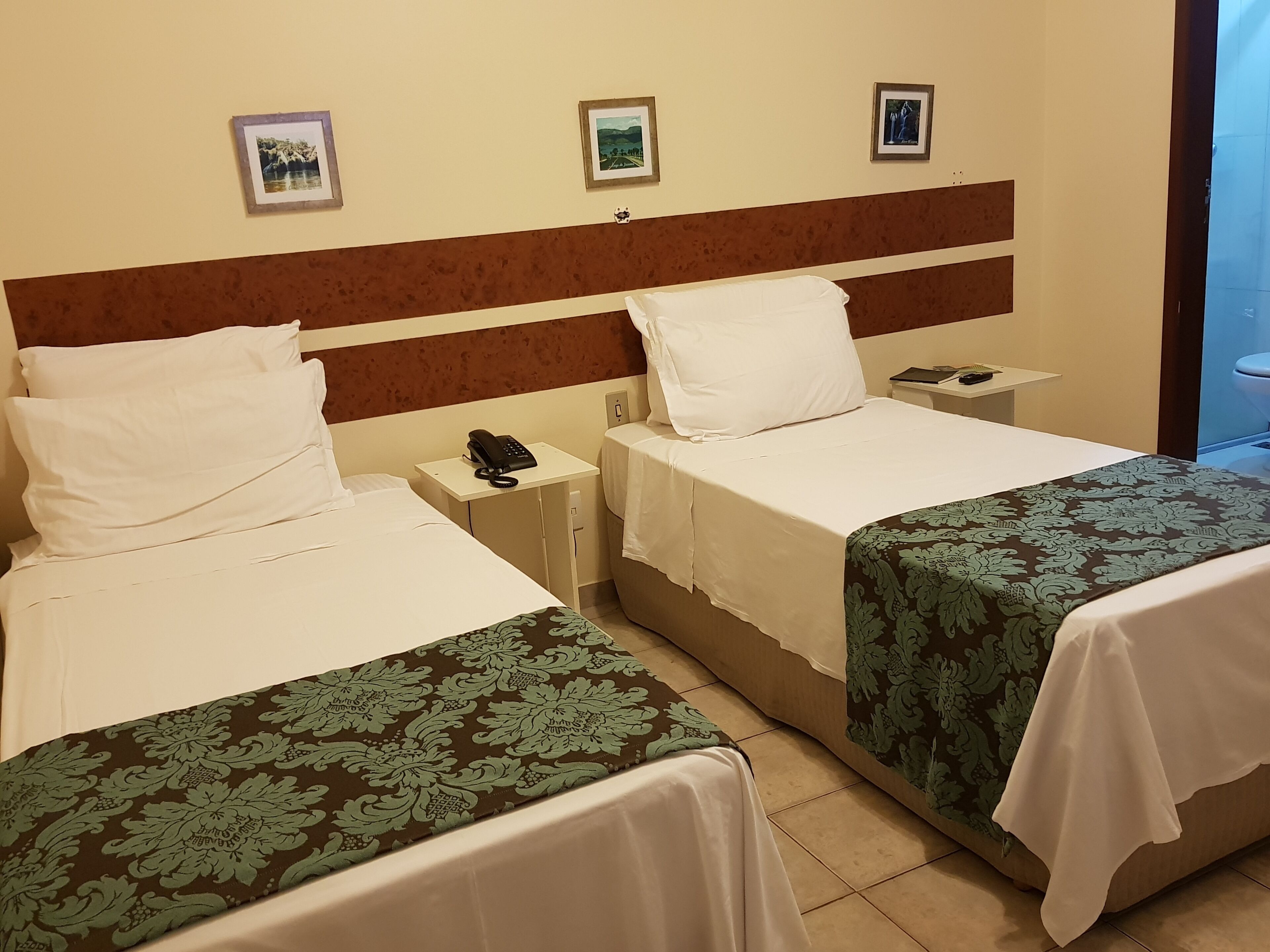 Quarto de hotel com duas camas, mesa de cabeceira com telefone e quadros nas paredes.