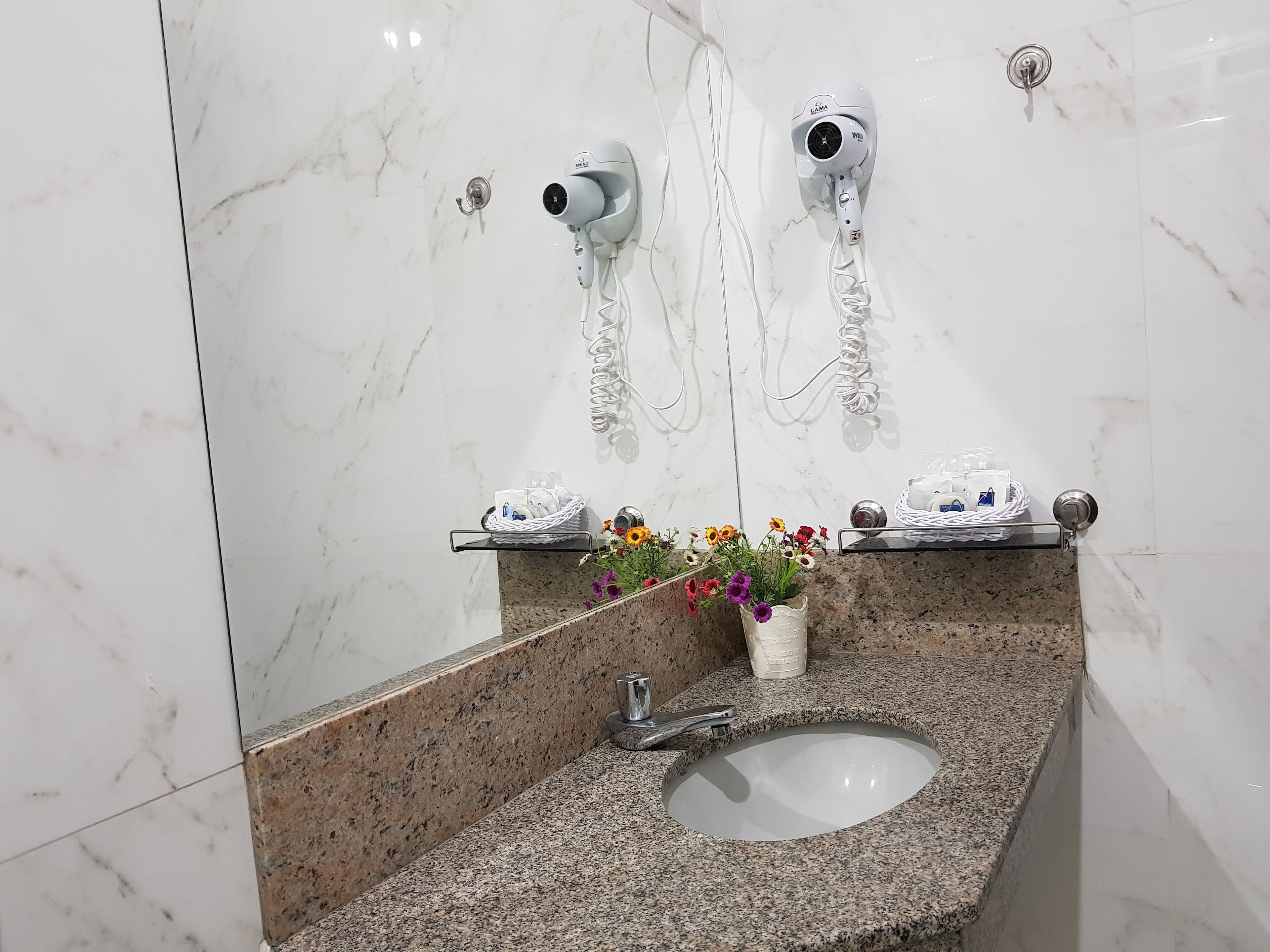 A bathroom with a marble wall, a granite countertop, a sink, a mirror, a hairdryer, and a vase with flowers.