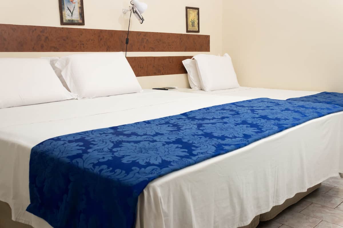 A neatly made bed with white linens and a blue floral bedspread, a wooden headboard, and a wall-mounted lamp.