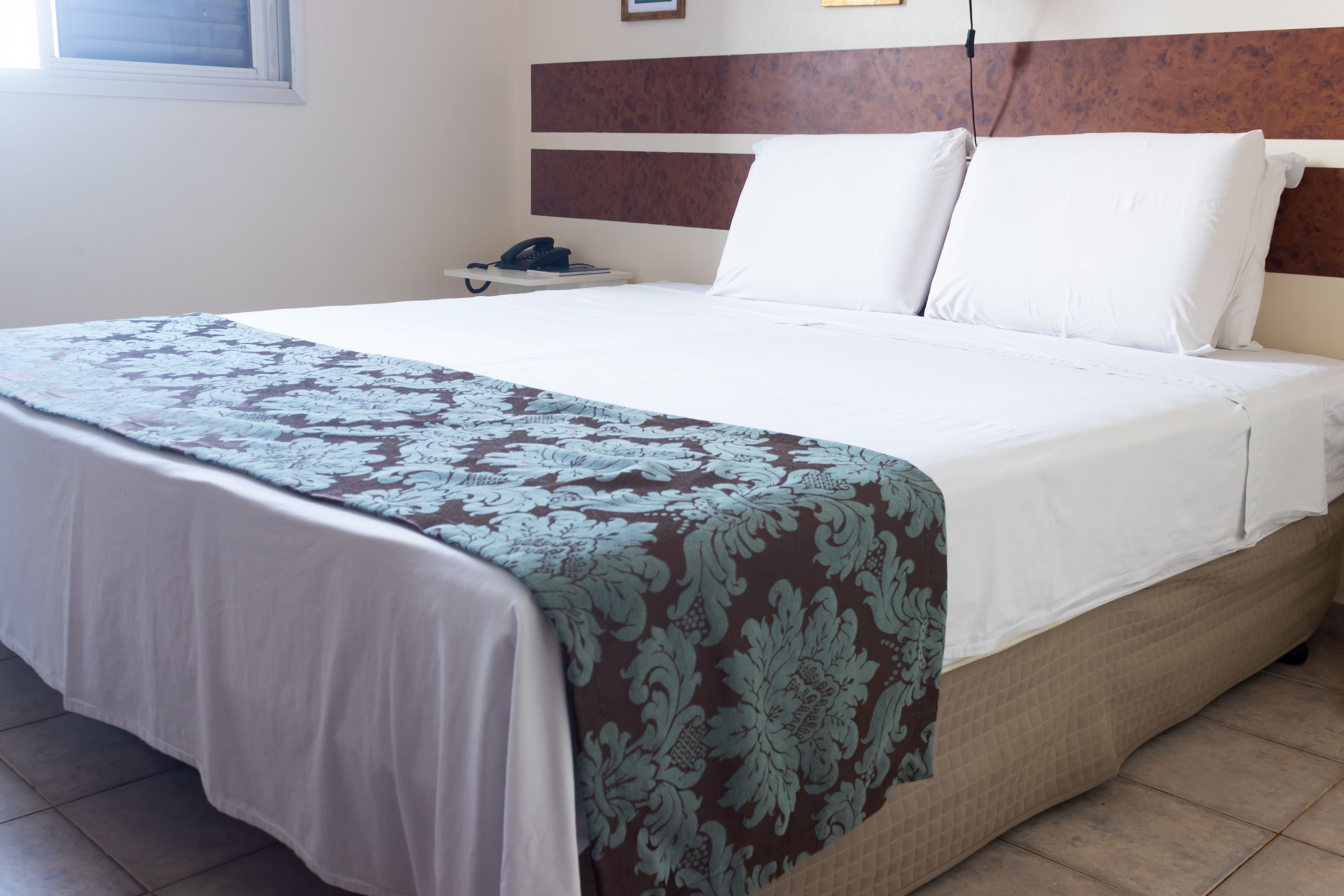 Una cama bien hecha con una colcha estampada, una mesita de noche con teléfono, y una lámpara de pared.