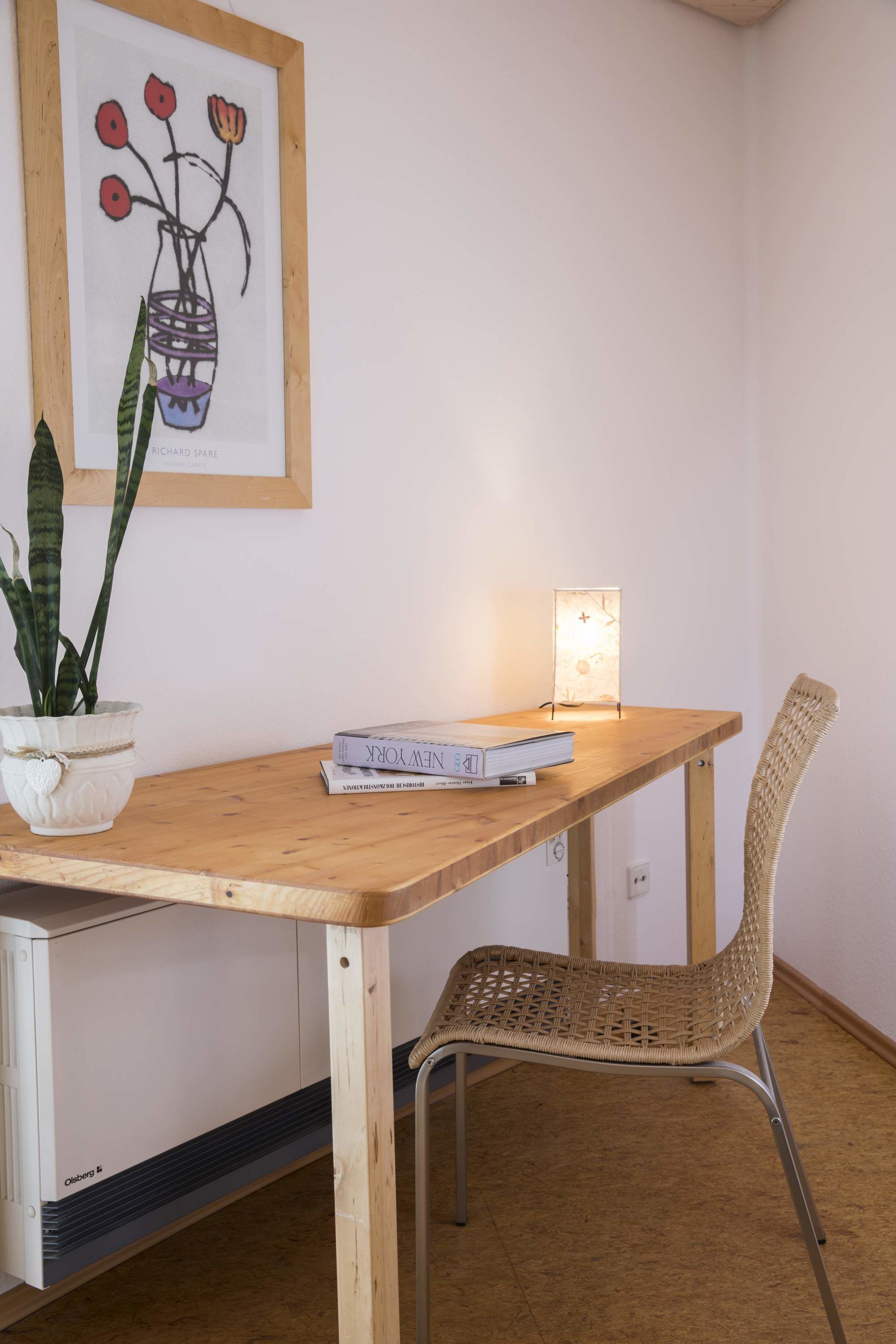 Een houten tafel met een rieten stoel, een bloempot en een lijst met kunstwerk aan de muur.