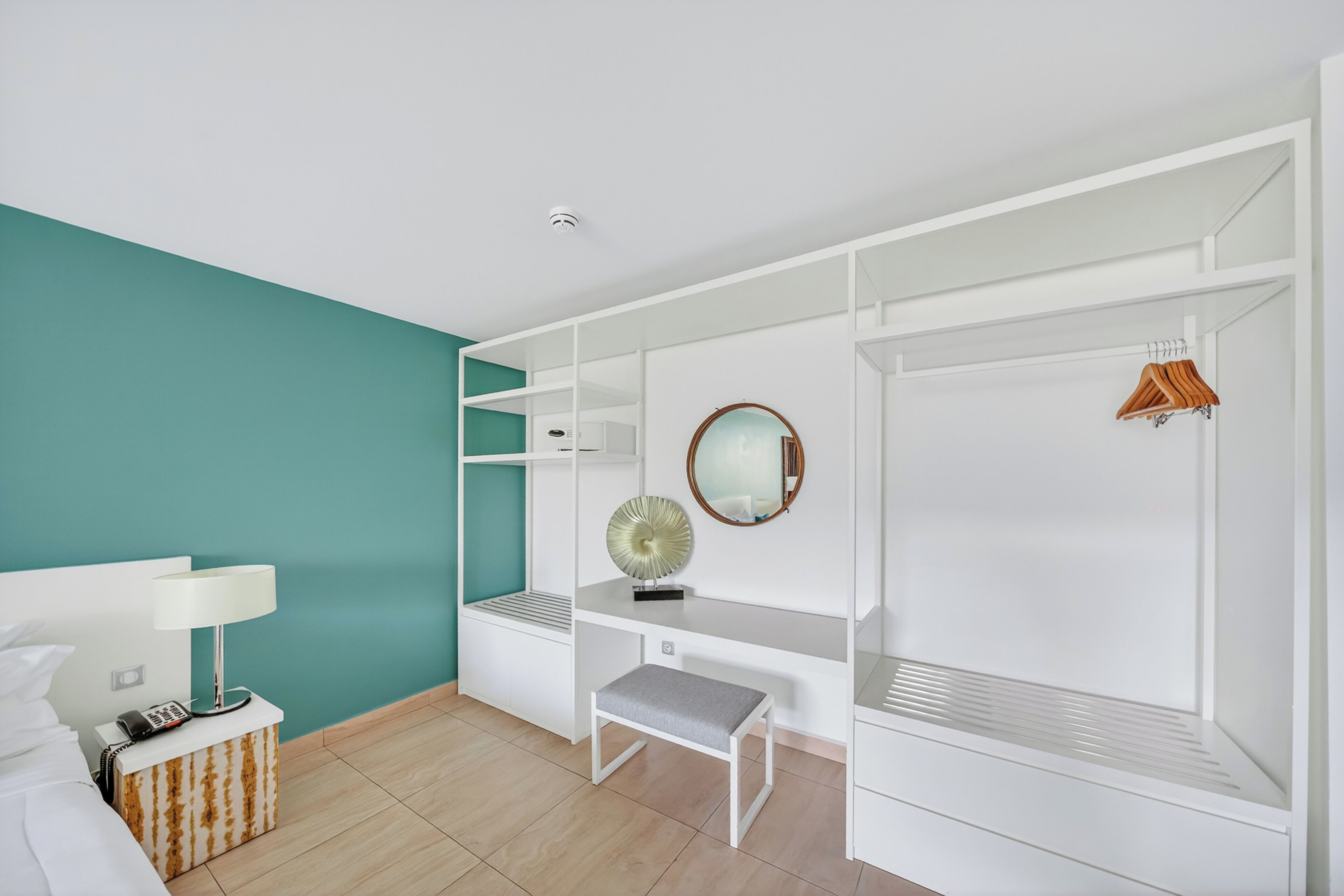 A modern bedroom with a teal accent wall, a white desk, a bench, a wardrobe, and a hanging clothes rack.