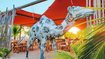 Una gran escultura blanca y texturizada de un animal parecido a un camello en un área arenosa al aire libre con muebles de madera y palmeras.