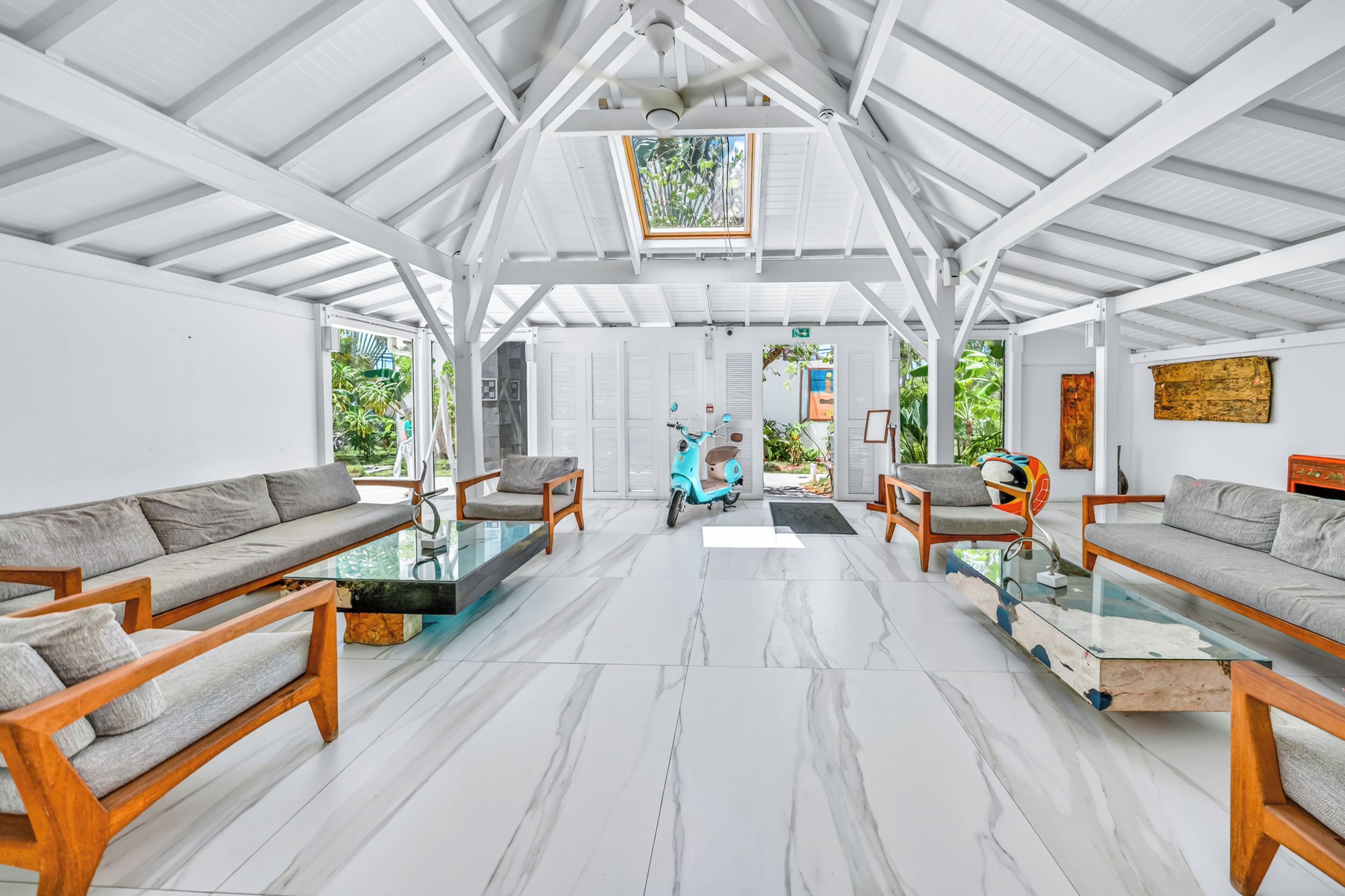 A spacious room with white walls, a high ceiling, and a skylight. There are two wooden sofas with cushions, a glass coffee table, and a teal scooter.