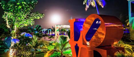 Área externa iluminada à noite, com uma grande escultura, palmeiras e um caminho bem iluminado.