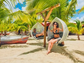 Dos personas descansando en sillones de mimbre bajo palmeras en una playa arenosa.