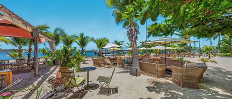 Un salón junto a la playa con asientos al aire libre, palmeras y vista al mar.