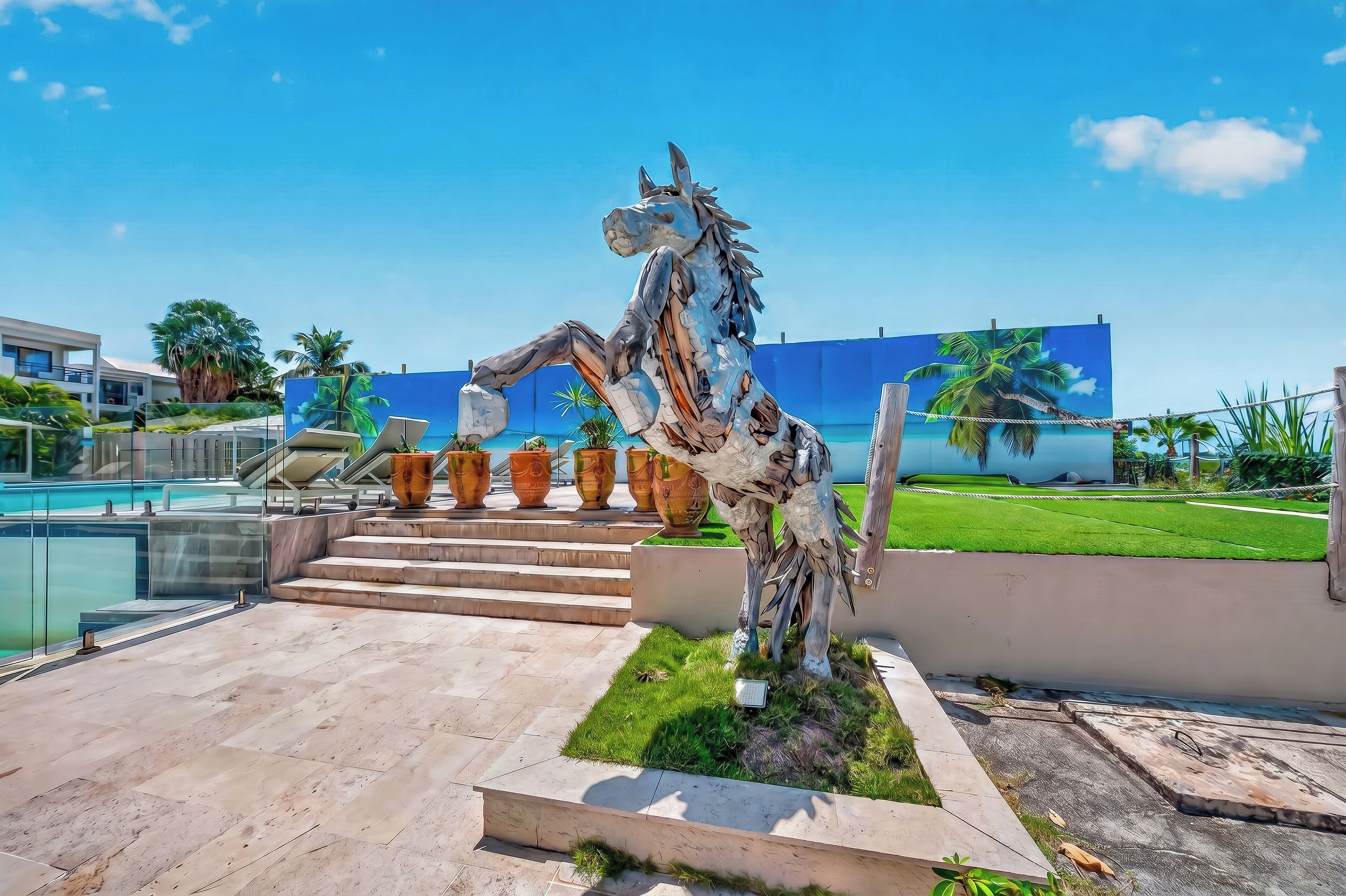 Un espacio exterior moderno con una escultura decorativa de un caballo, una piscina y un muro azul con palmeras.
