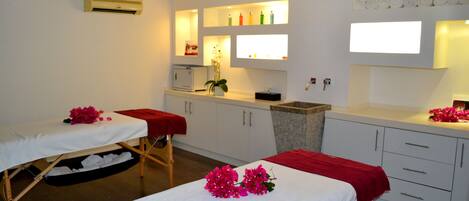 A spa room with two treatment beds, a small sink, and shelves with toiletries.