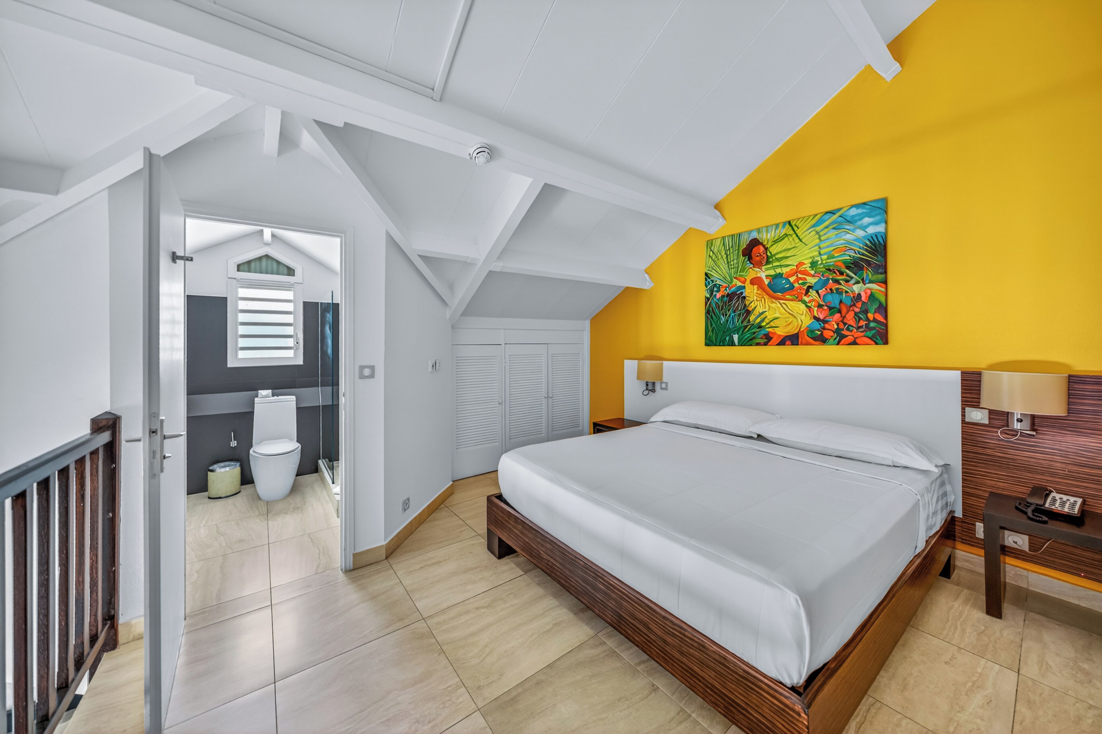 Un dormitorio con una cama, un cuadro, una mesita de noche y un baño visible a través de una puerta abierta.