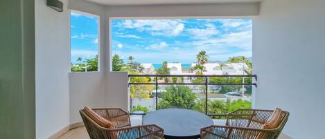 A balcony with a round table and wicker chairs, overlooking a residential area with houses and greenery.