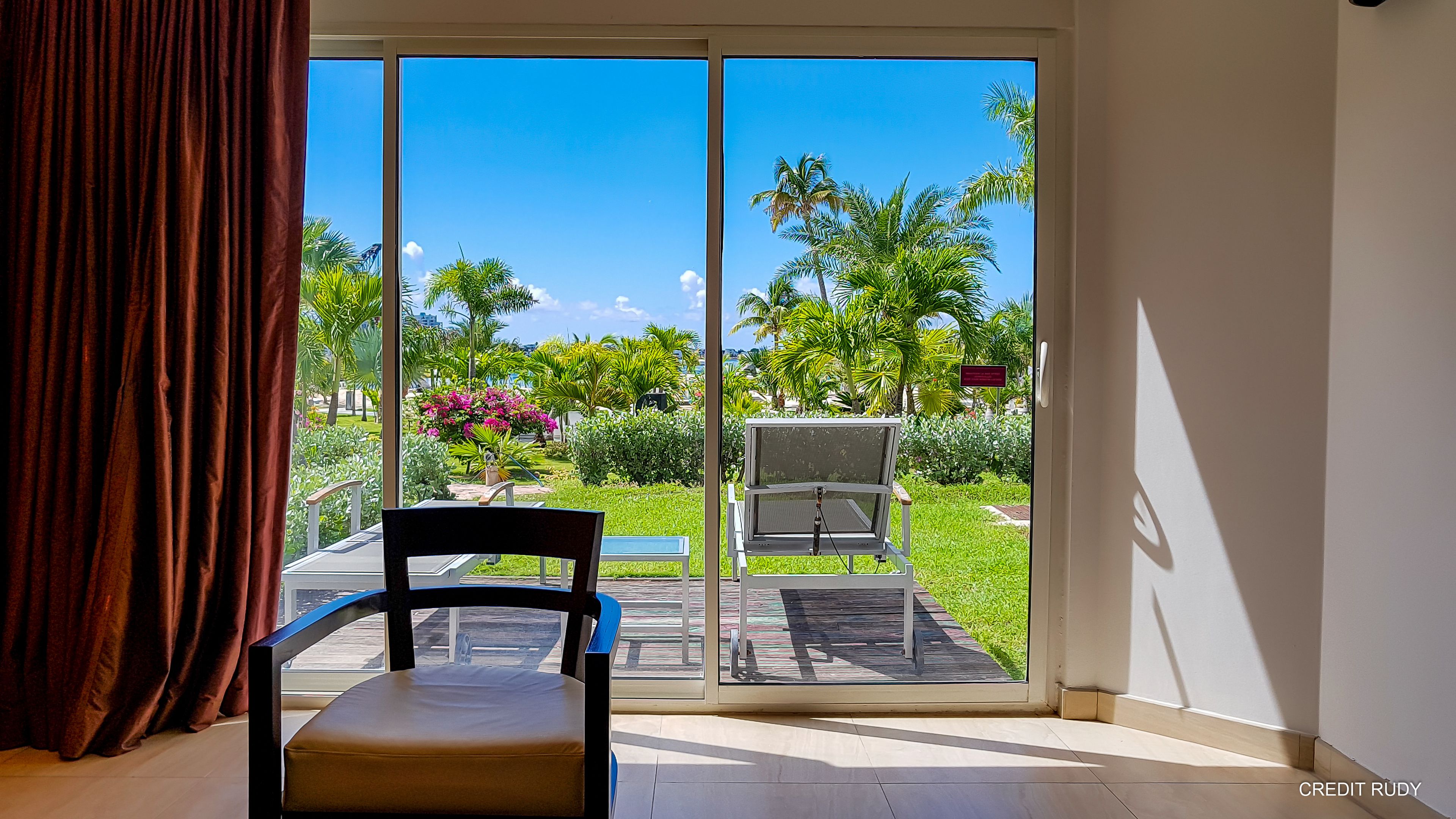 A room with a sliding glass door leading to a garden with palm trees and a bench.